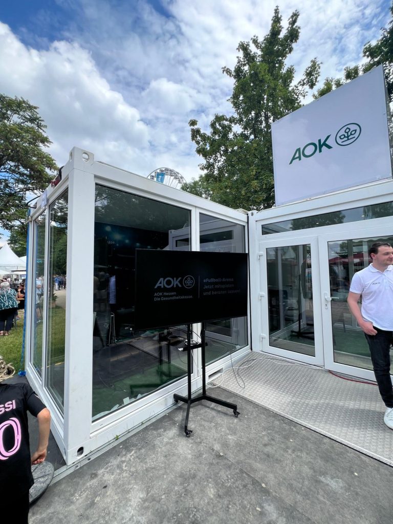 Hessentag AOK #6 Der Blick auf die eFußball-Arena und den Gameplay-Bildschirm im verglasten Container beim Hessentag im Auftrag der AOK.